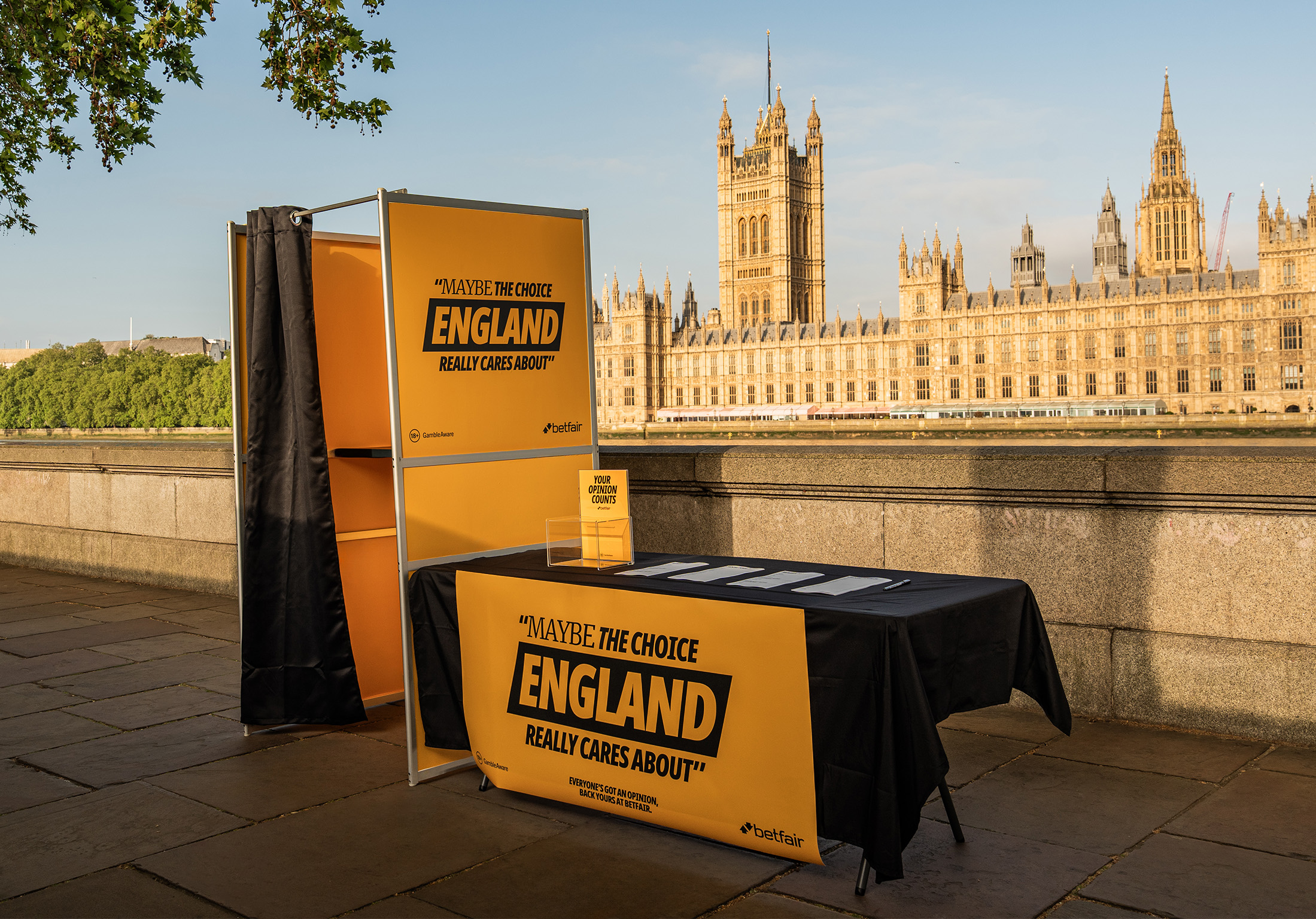 A Betfair-branded polling booth and table set up on the South Bank of the Thames, with the Houses of Parliament and Victoria Tower in the background. The booth signage reads: 'Maybe the choice England really cares about.' A small sign on the table says 'Your opinion counts.'