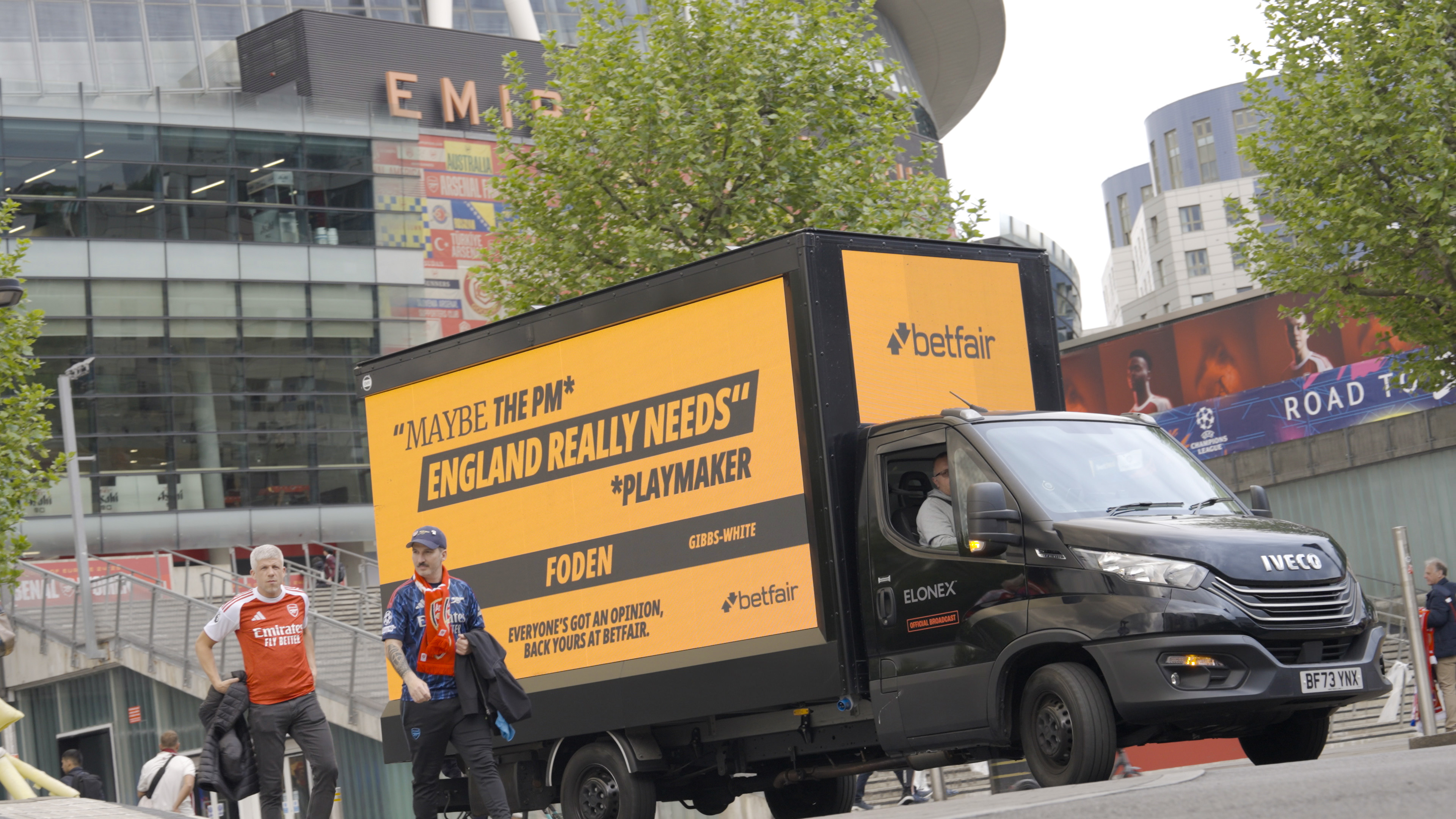 A black Betfair-branded ad van parked outside the Emirates Stadium, Arsenal's ground. The yellow LED side reads 'Maybe the PM* England really needs' with an asterisk note '*Playmaker.' Two names below: FODEN and GIBBS-WHITE. A pair of Arsenal-shirted fans, one a Mikel Arteta lookalike, stand beside the van.