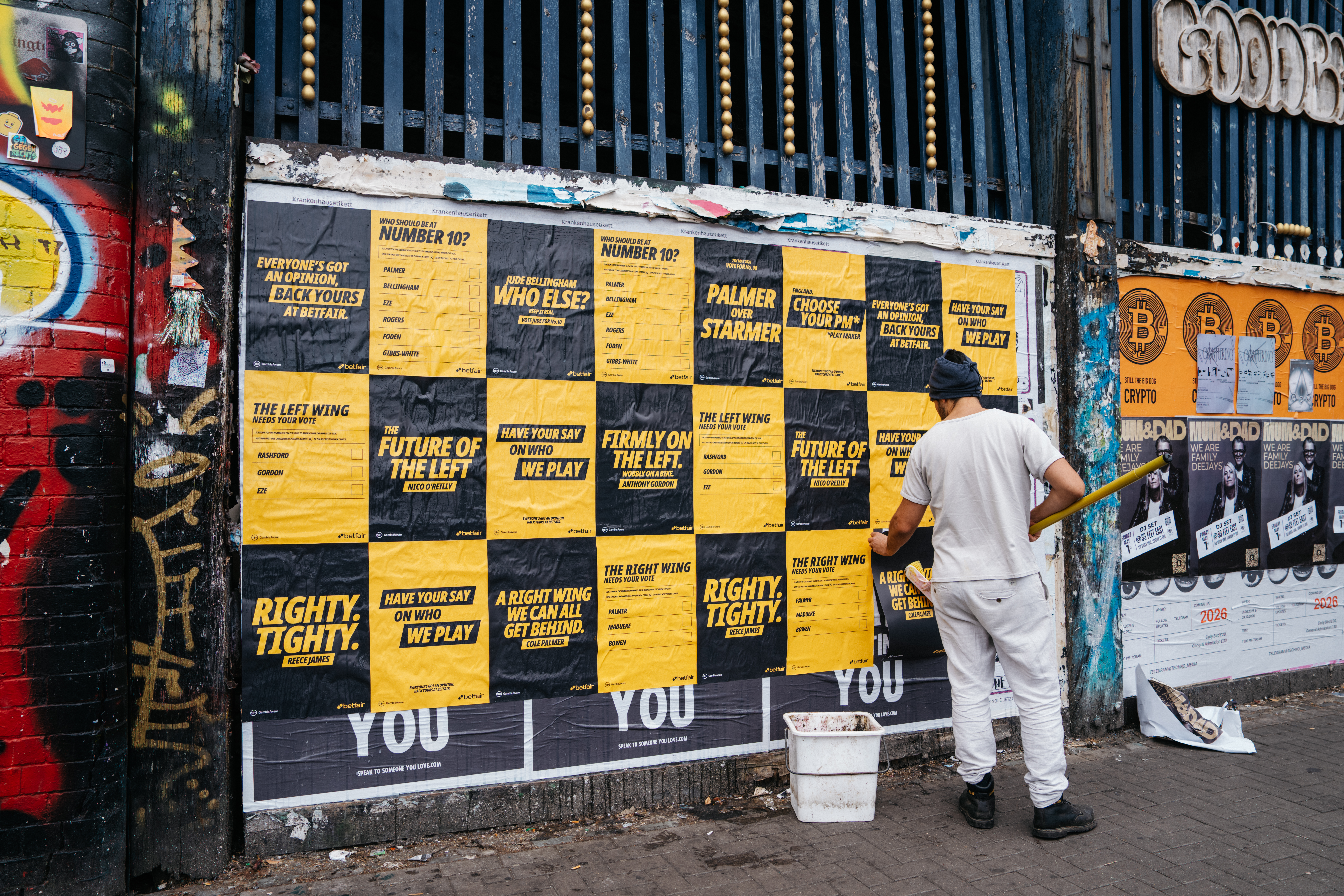 A fly-posterer pastes up a wall of black-and-yellow Betfair posters in an urban street setting. The posters carry various 'Maybe' lines: 'Who should be at Number 10?', 'Jude Bellingham who else?', 'Palmer over Starmer', 'Choose your PM*Play Maker', 'Righty. Tighty. Reece James', 'The future of the left. Nico O'Reilly', 'Firmly on the left. Wobbly on a bike. Anthony Gordon'.