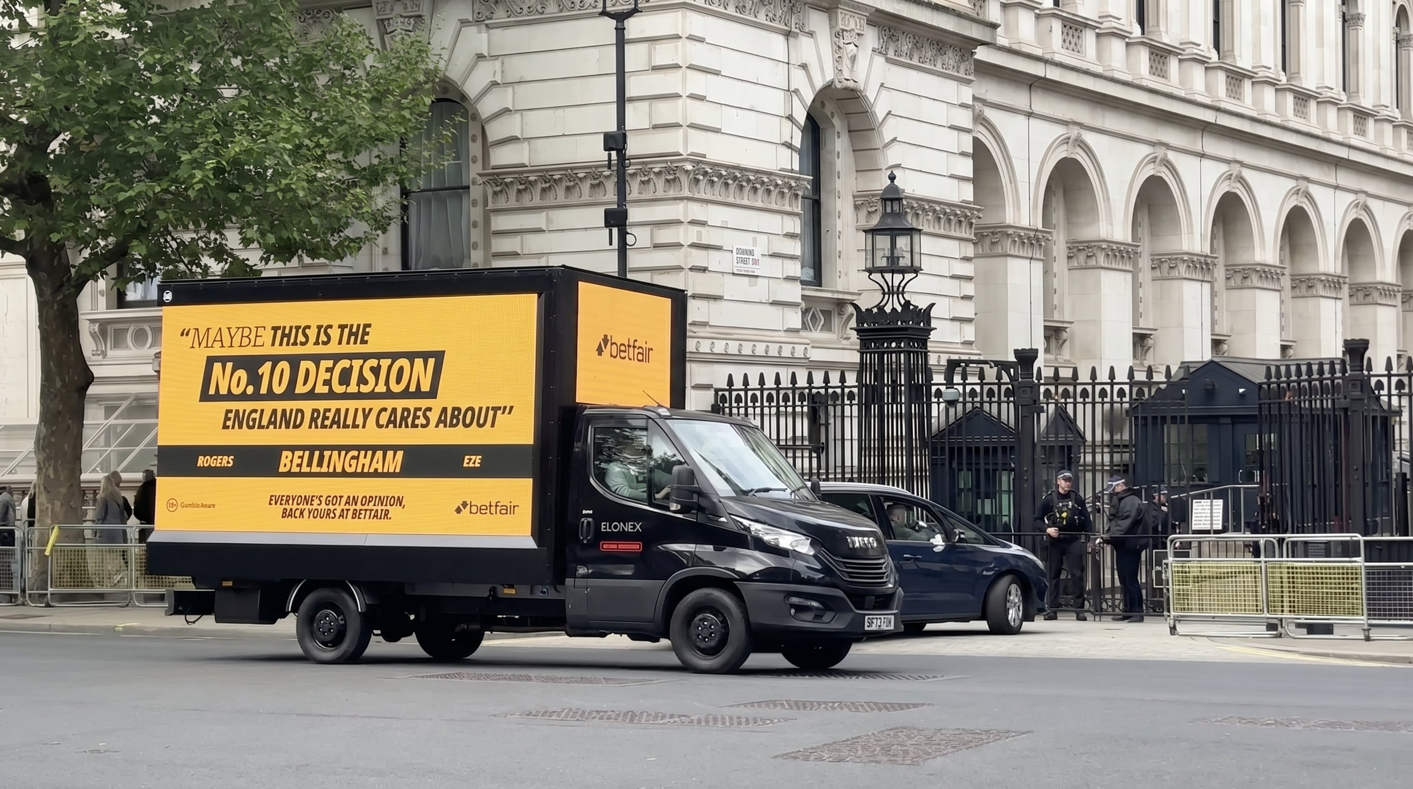 A black Betfair-branded media van parked outside the Downing Street gates in central London. The yellow LED side reads: 'Maybe this is the No.10 decision England really cares about.' Underneath, three candidate names: ROGERS, BELLINGHAM, EZE. Footer: 'Everyone's got an opinion, back yours at Betfair.' Police officers and a Downing Street sign visible to the right.