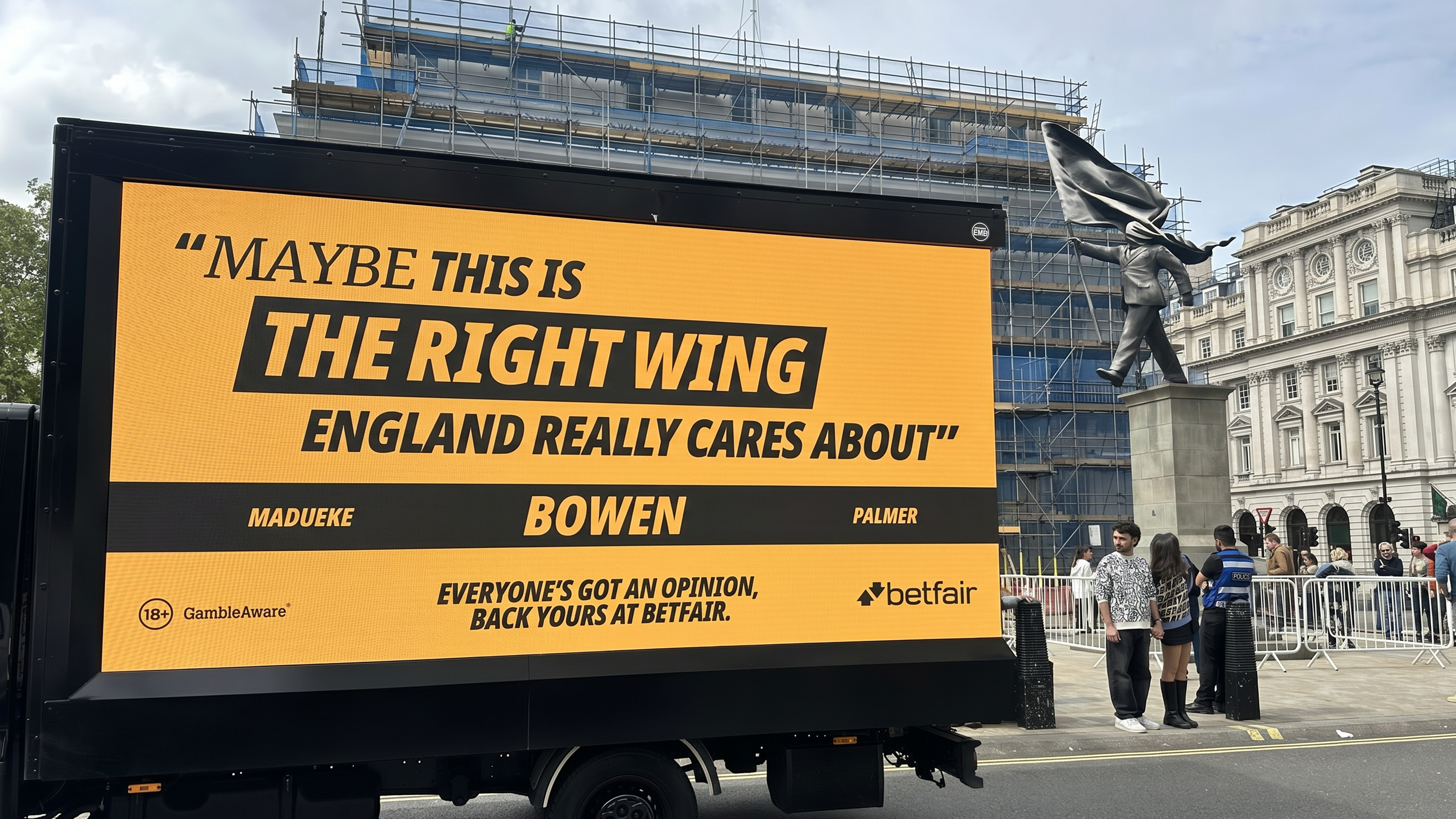 A black Betfair-branded ad van with a yellow LED side reading 'Maybe this is the right wing England really cares about.' Three names below: MADUEKE, BOWEN, PALMER. Footer: 'Everyone's got an opinion, back yours at Betfair.' A statue of a figure carrying a white flag is visible in the background.