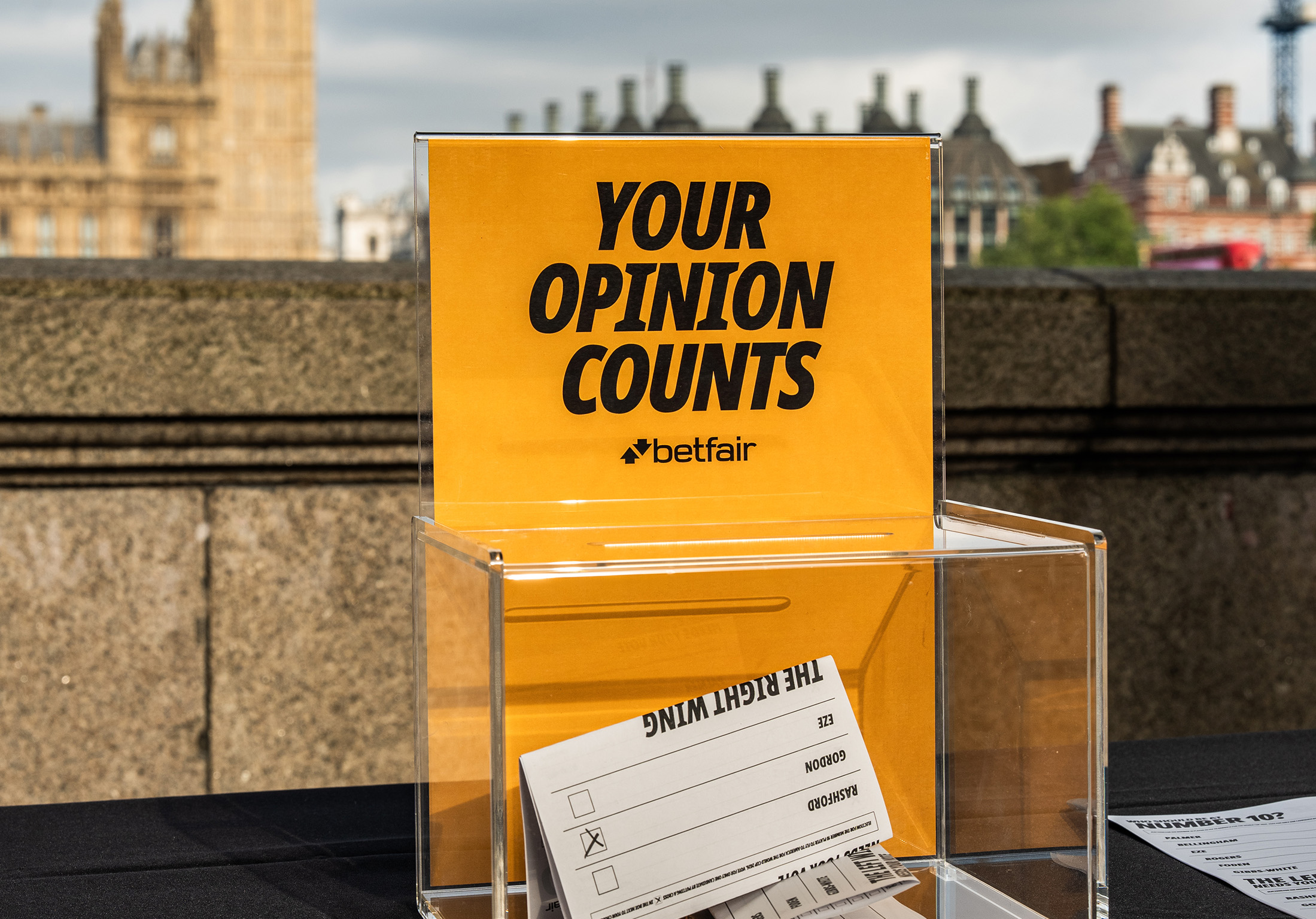 A close-up of a clear acrylic Betfair ballot box with an amber yellow sign on top reading 'Your opinion counts' alongside the Betfair logo. A completed paper ballot card titled 'The Right Wing' is visible inside and beside the box. Parliament's stonework is softly out-of-focus in the background.