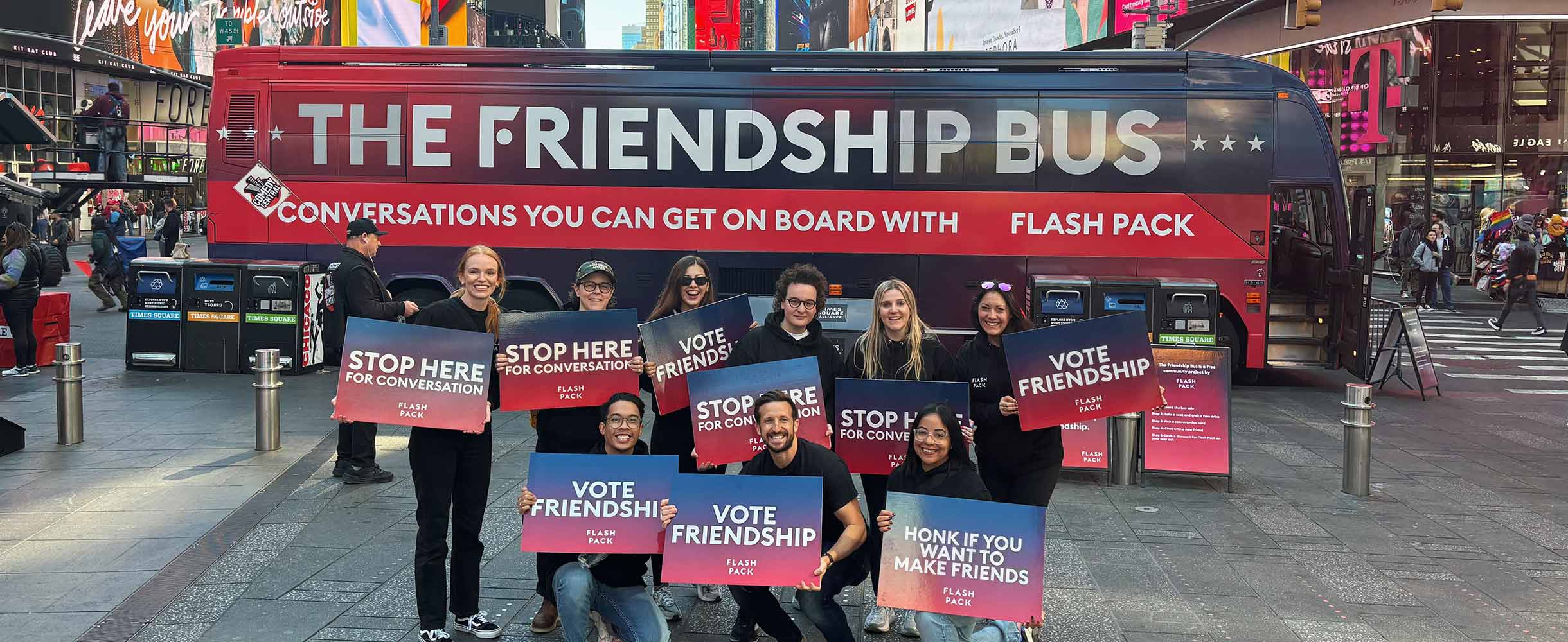 The Friendship Bus parked in Times Square, New York. Team members hold signs reading 'STOP HERE FOR CONVERSATION' and 'VOTE FRIENDSHIP' in coral-to-blue gradient against the bus livery.