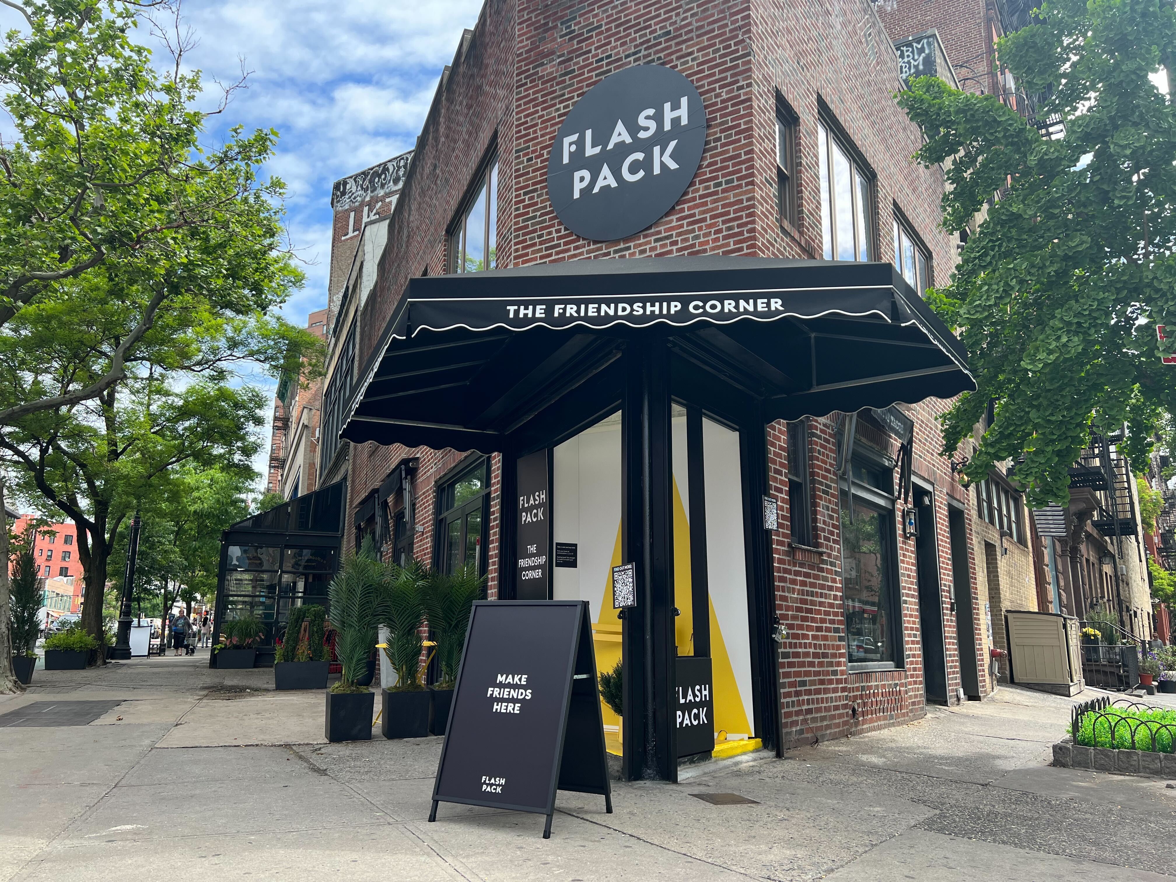 The Friendship Corner pop-up storefront in the West Village, New York. A black awning reading 'THE FRIENDSHIP CORNER' over a Flash Pack-branded interior, with a sandwich board outside reading 'MAKE FRIENDS HERE'.